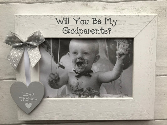 Image shows wooden photo frame for god parents, consists of wooden heart with name of god child on, attached to white ribbon and a dotted bow.