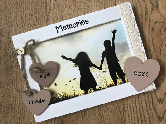 Image shows lace memories photo frame, consists of two hanging hearts for names and a small wooden heart above, on the same is lace ribbon and another heart for year.