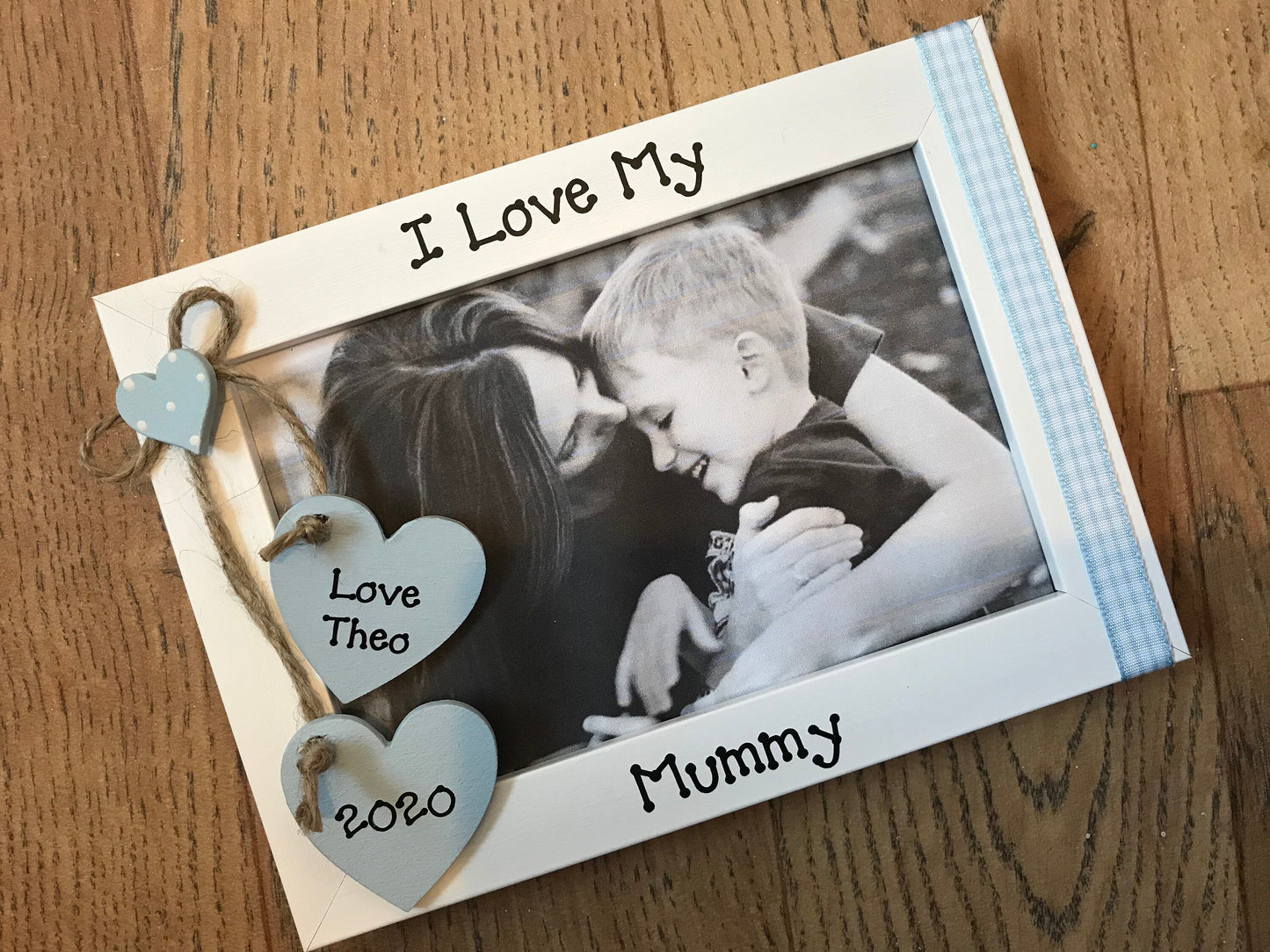 Image shows i love my mummy photo frame, attached is two hanging wooden hearts with a name and date, on the opposite side there is gingham ribbon attached.